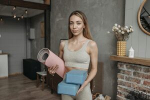 kobieta ubrana w beżowy strój sportowy i trzymająca różową matę do ćwiczeń oraz niebieksie klocki do pilatesu