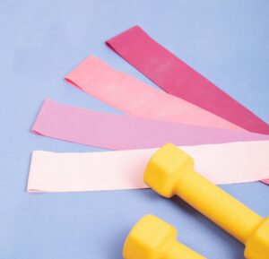 gumy do pilatesu w czterech odcieniach różowego obok żółtych hantli na lawendowym tle