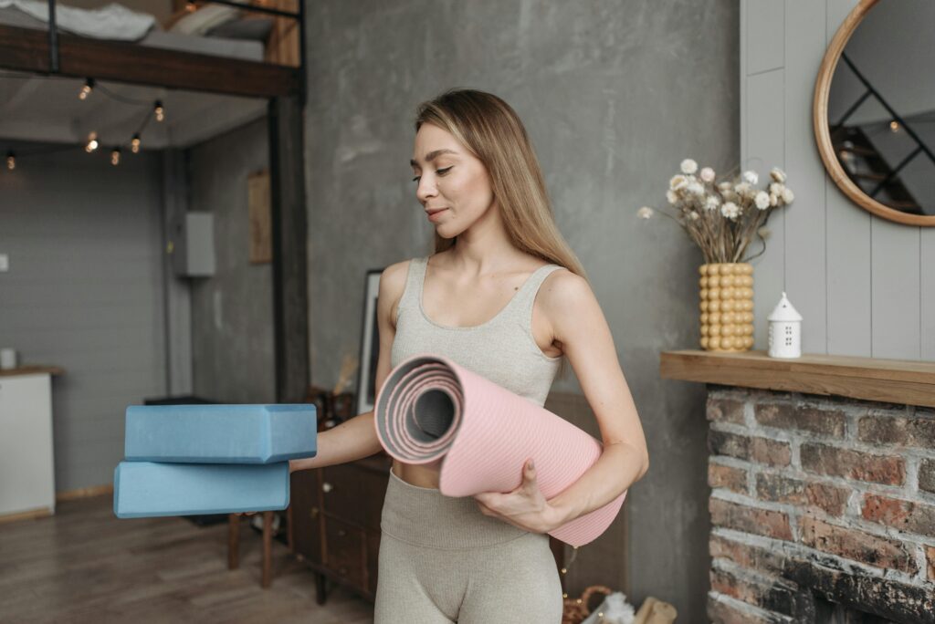 kobieta w blond włosach trzymająca niebieskie klocki do pilatesu i różową matę w drugiej ręce, zerkająca na te akcesoria do pilatesu z błogim uśmiechem 